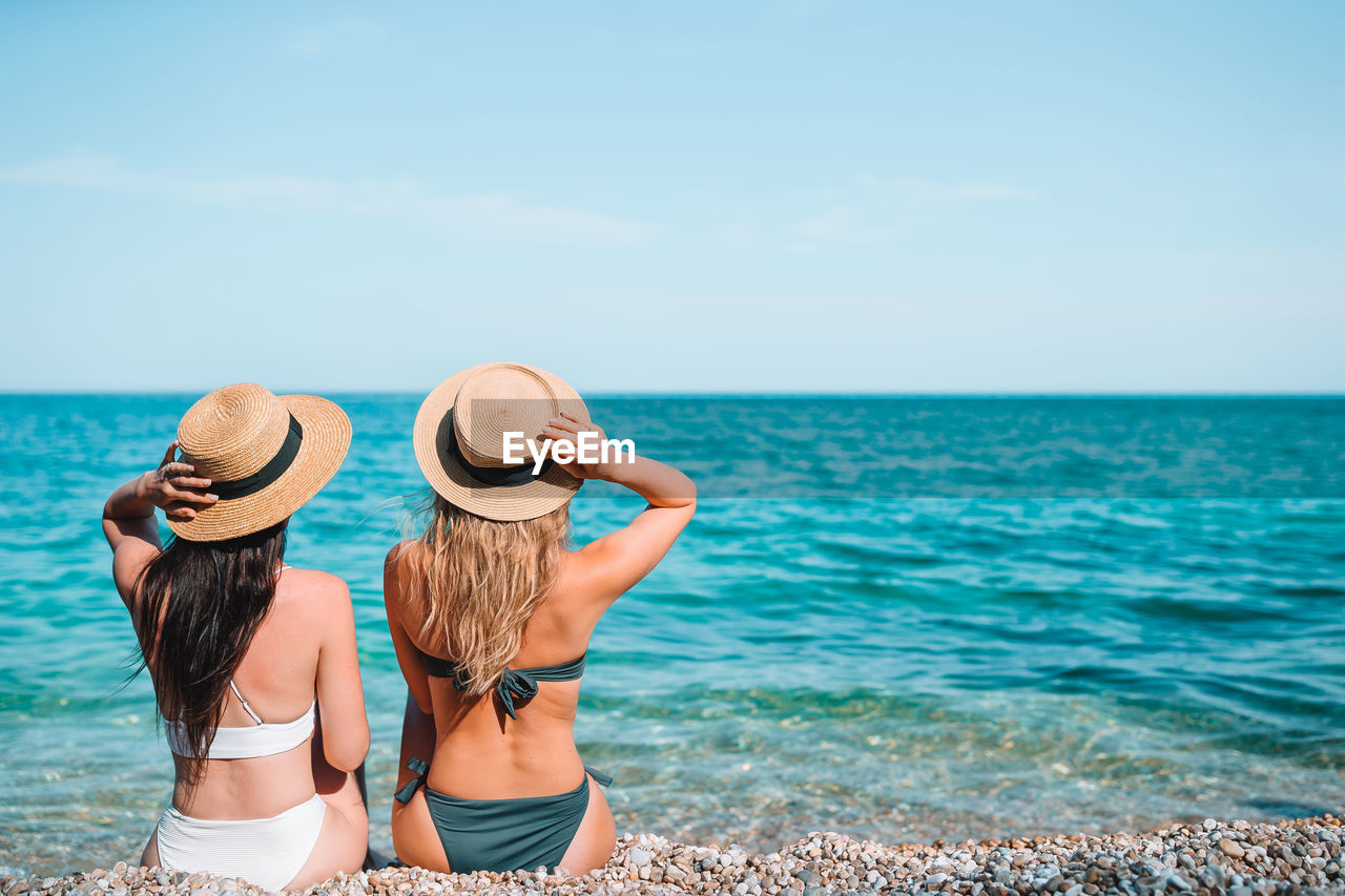 Rear view of women wearing hat sitting on beach against sea