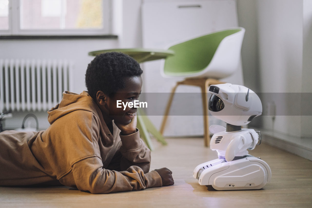 Smiling boy talking with ai robot while lying on floor in innovation lab