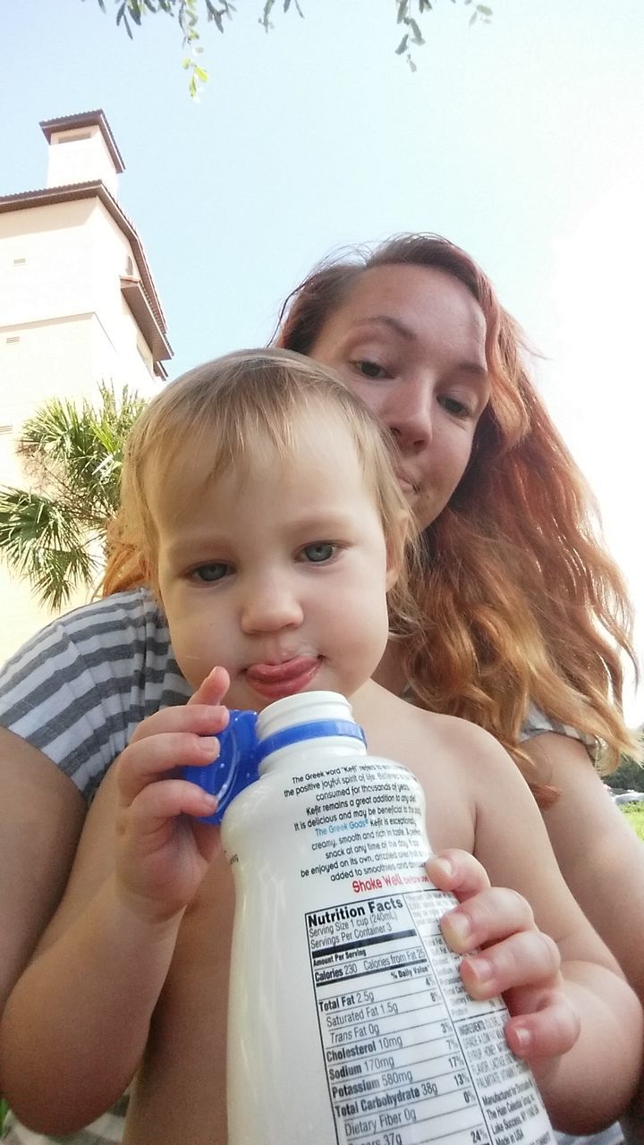 Low angle portrait of baby with mother drinking milk against clear sky