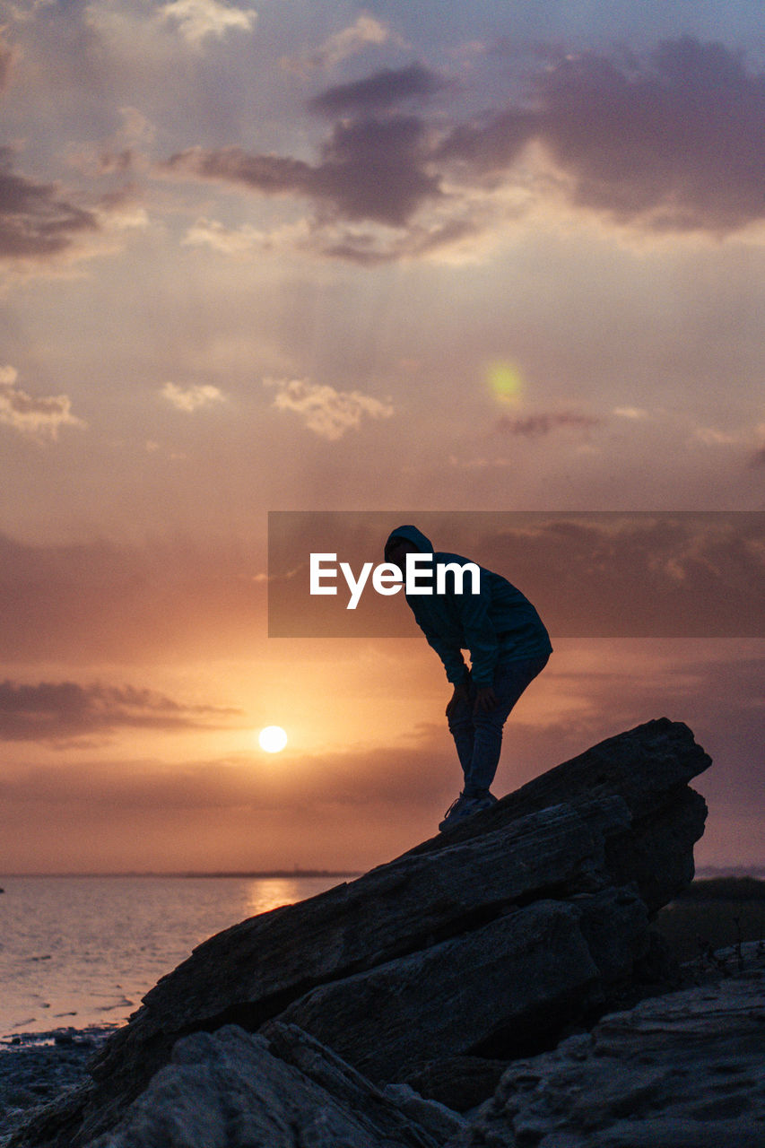 Man standing on rock by sea against sky during sunset