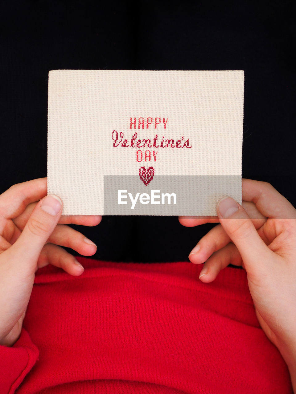 Child in a red sweater holding a handmade postcard with embroidery happy valentine's day.