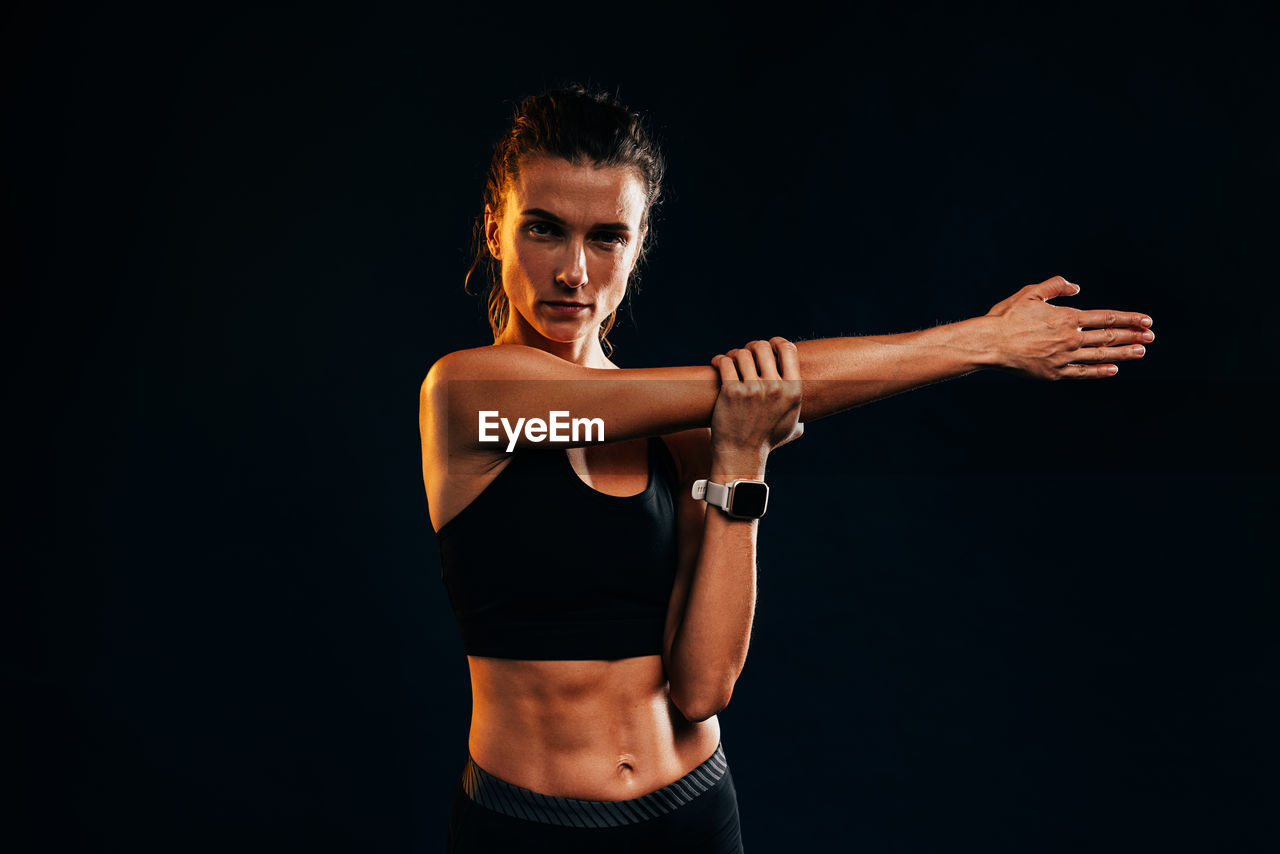 portrait of young woman exercising with arms raised standing against black background