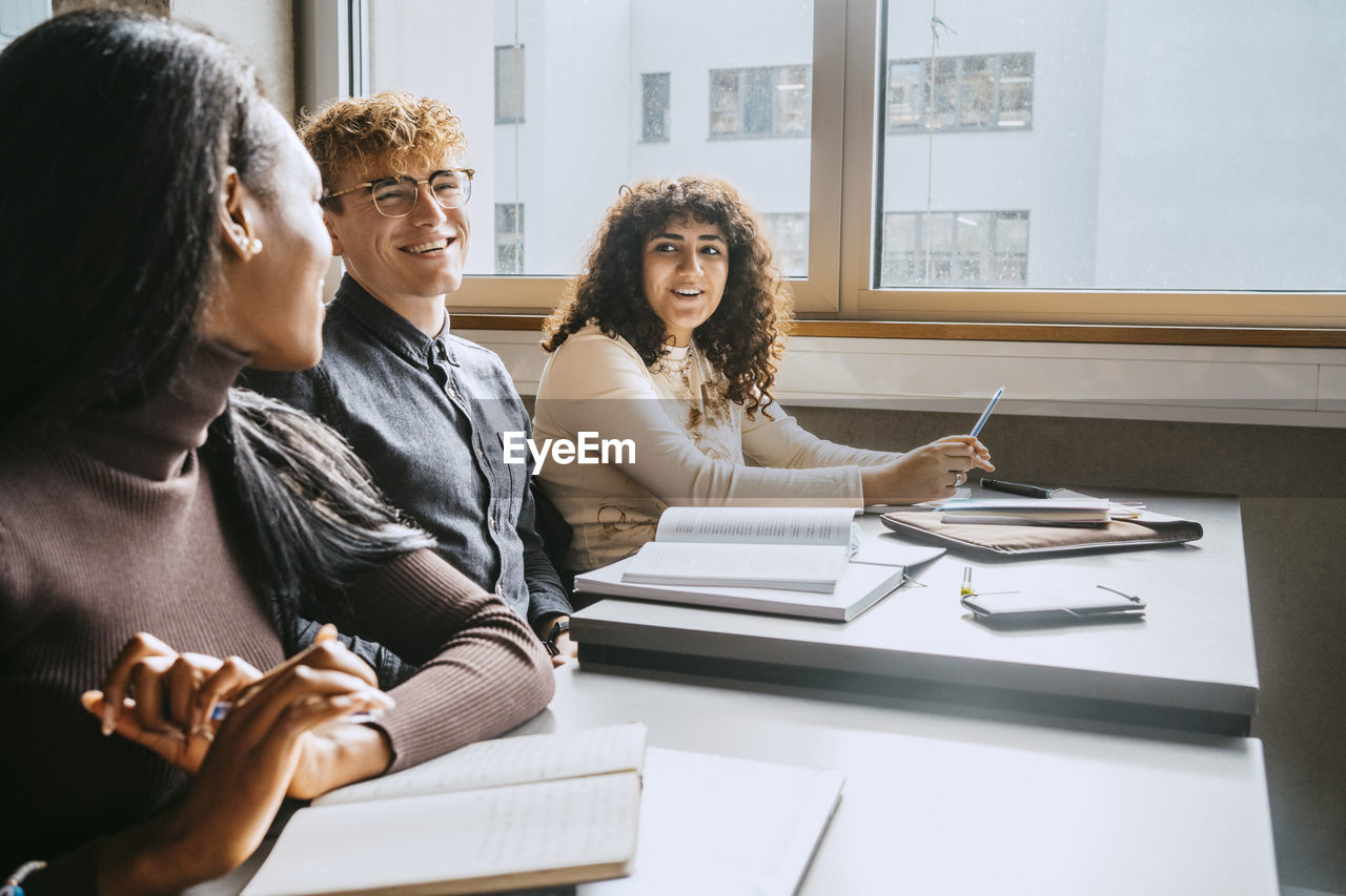 Smiling young multiracial friends sitting together at desk in university classroom