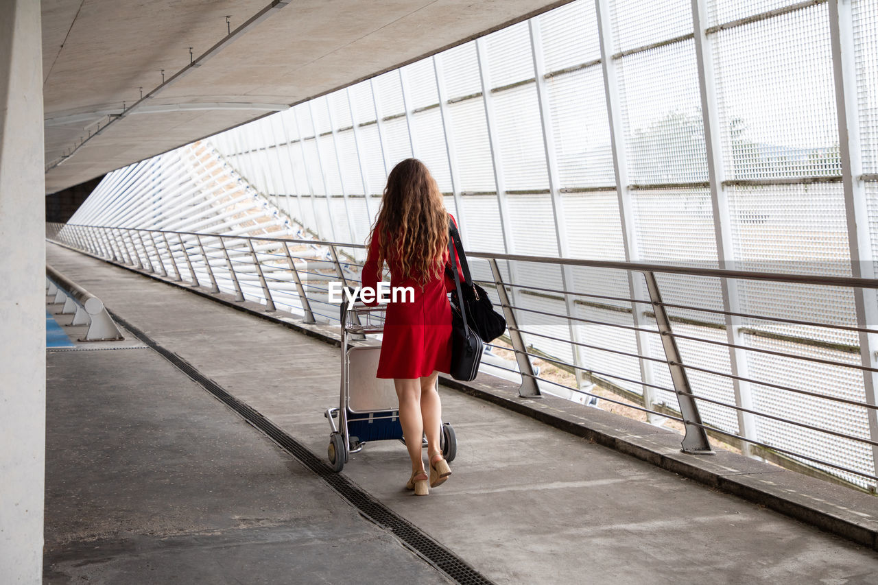 Back view of traveling female walking in airport with baggage trolley and waiting for flight