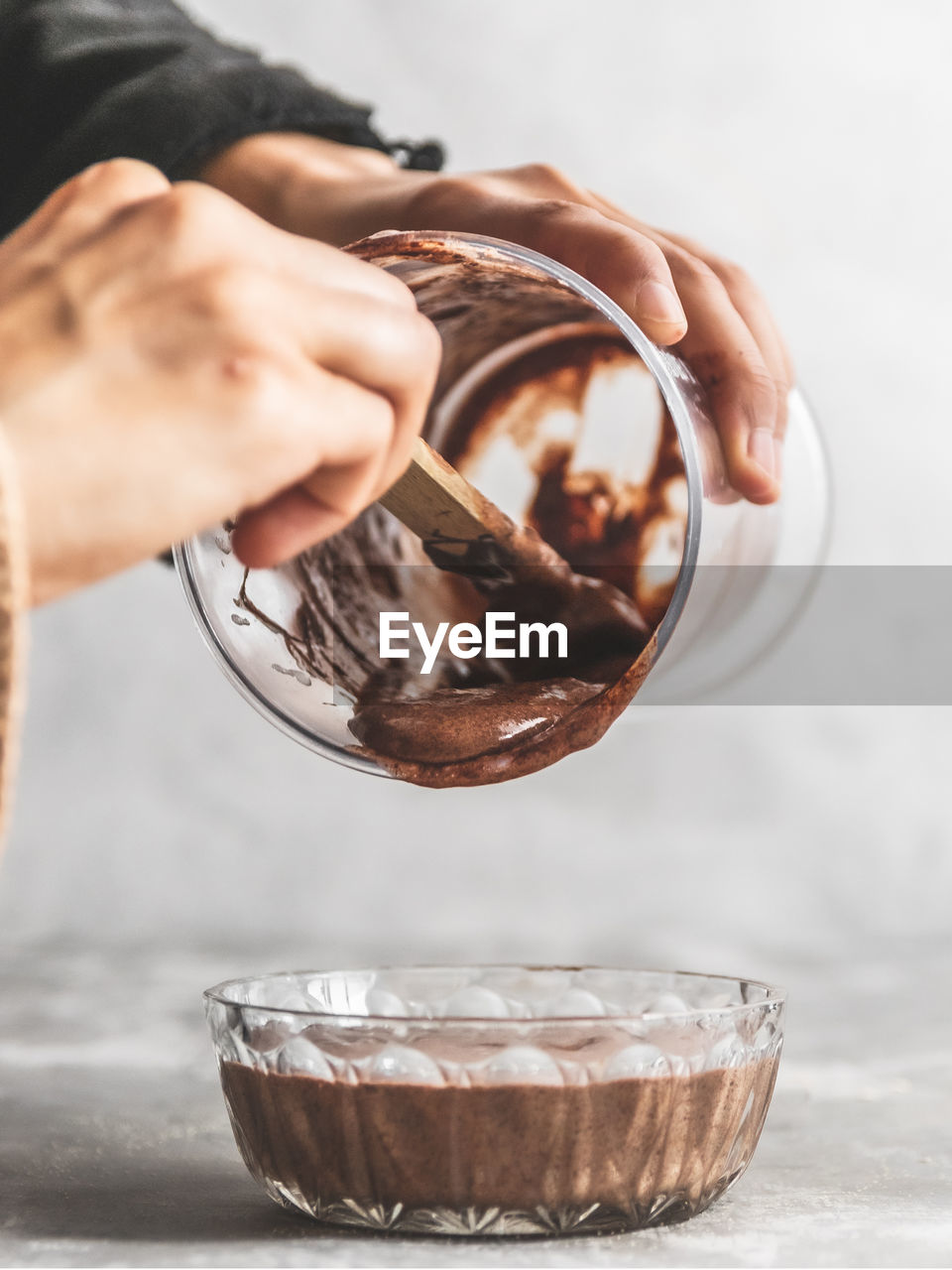 Close-up of hand holding bowl of chocolate mousse batter pouring into glass bowl