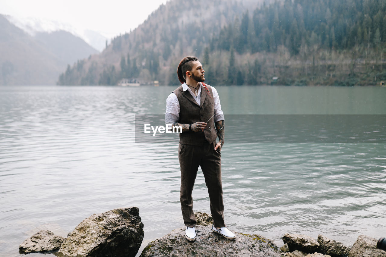 A man a groom a hipster with dreadlocks in a wedding suit stands by sea and mountains in nature