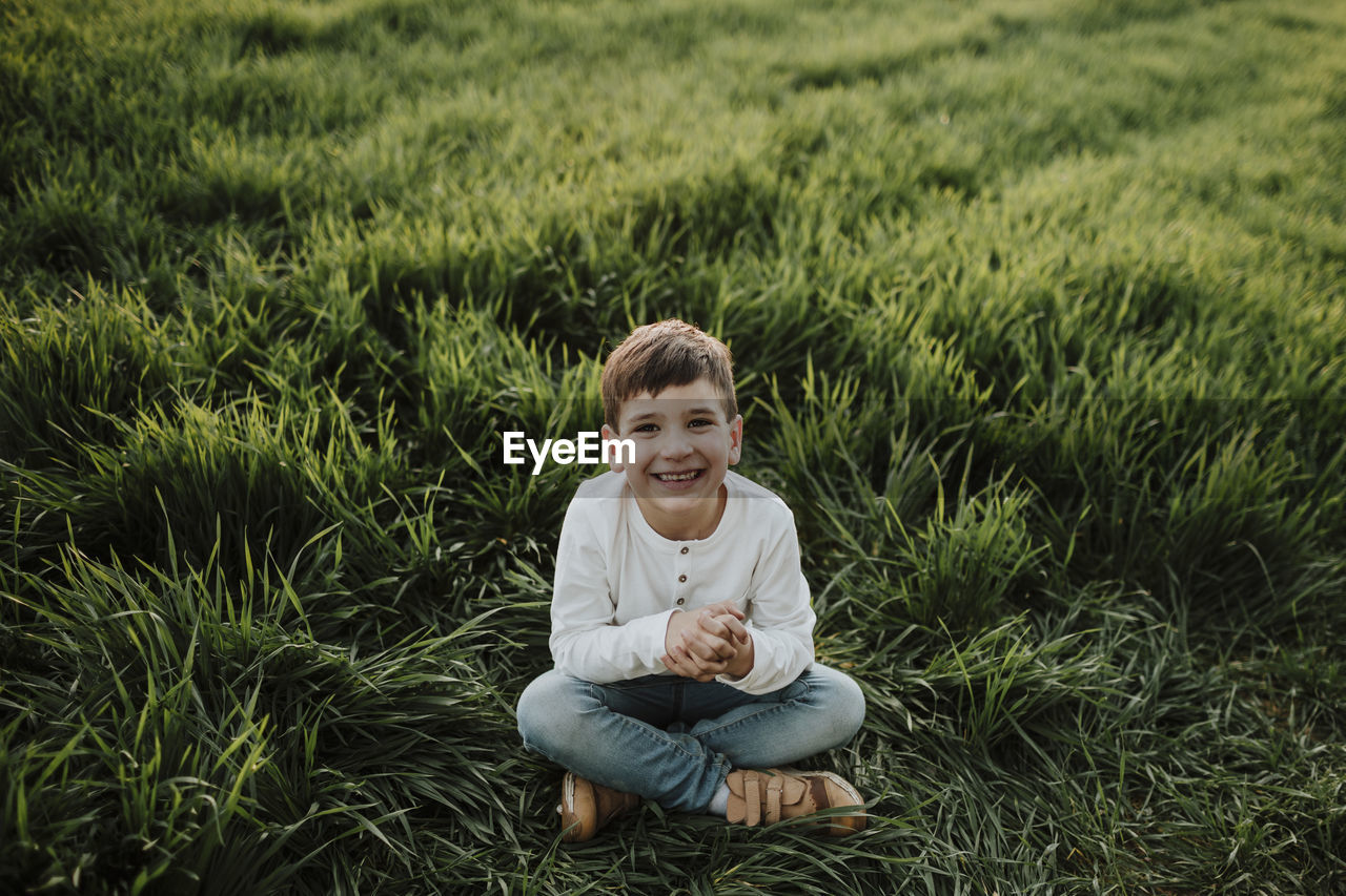 Cheerful boy with hands clasped sitting at grassy field