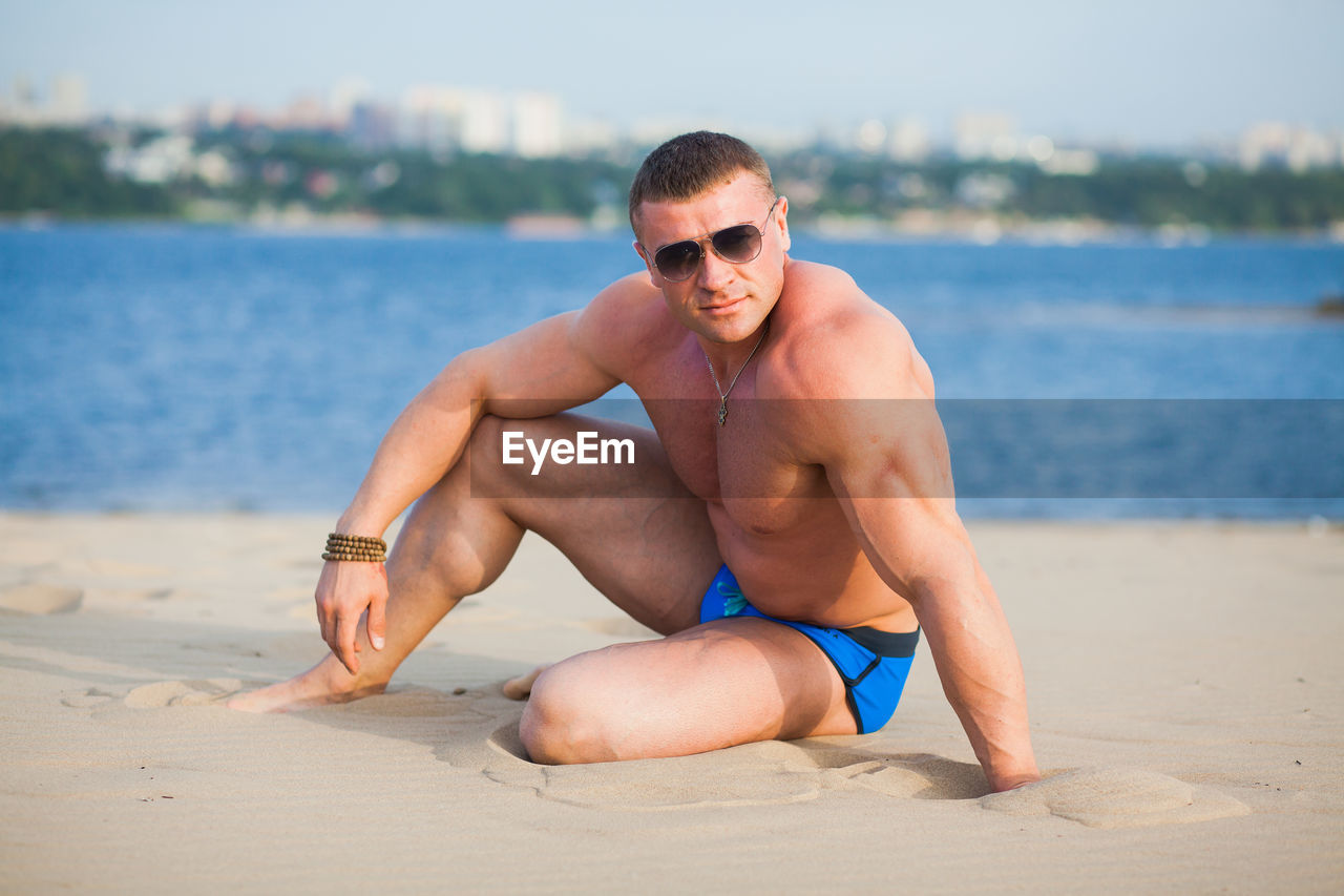 Portrait of shirtless bodybuilder while standing on beach against bay of water