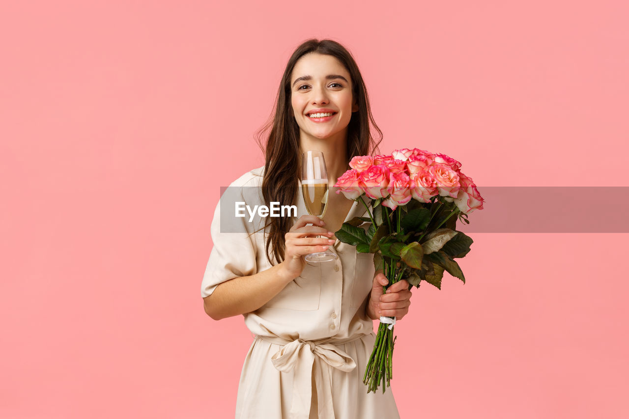 PORTRAIT OF HAPPY YOUNG WOMAN HOLDING PINK FLOWER AGAINST GRAY BACKGROUND