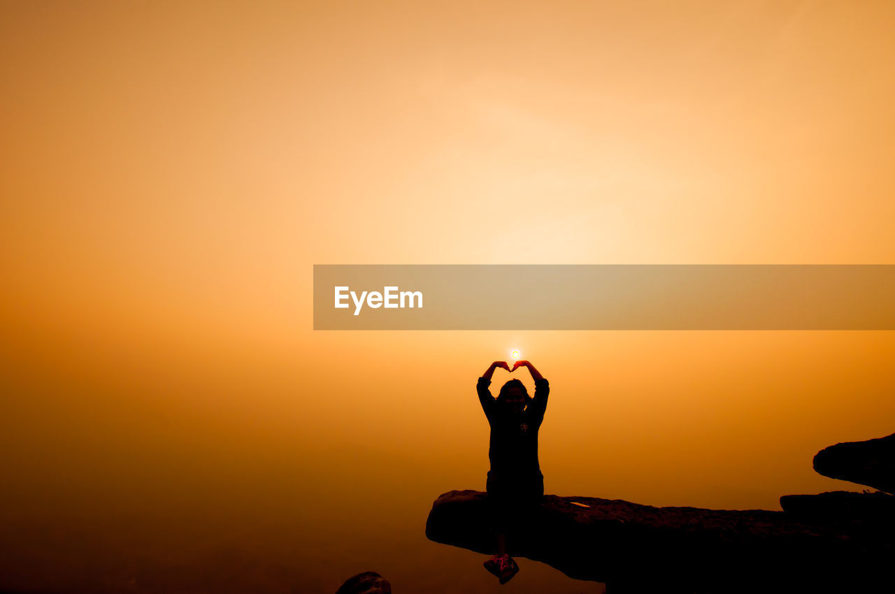 Silhouette person making heart shape on cliff against sky during sunset