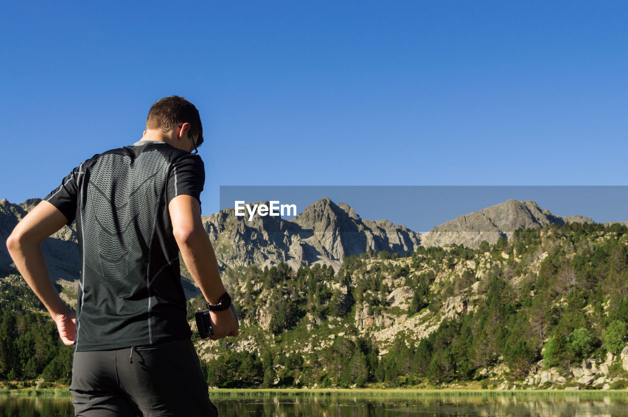 REAR VIEW OF MAN STANDING ON MOUNTAIN AGAINST CLEAR SKY