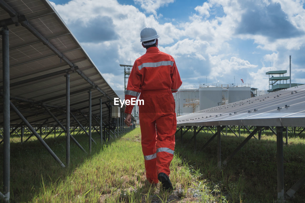 Rear view of worker wearing reflective clothing while standing on solar panel against sky