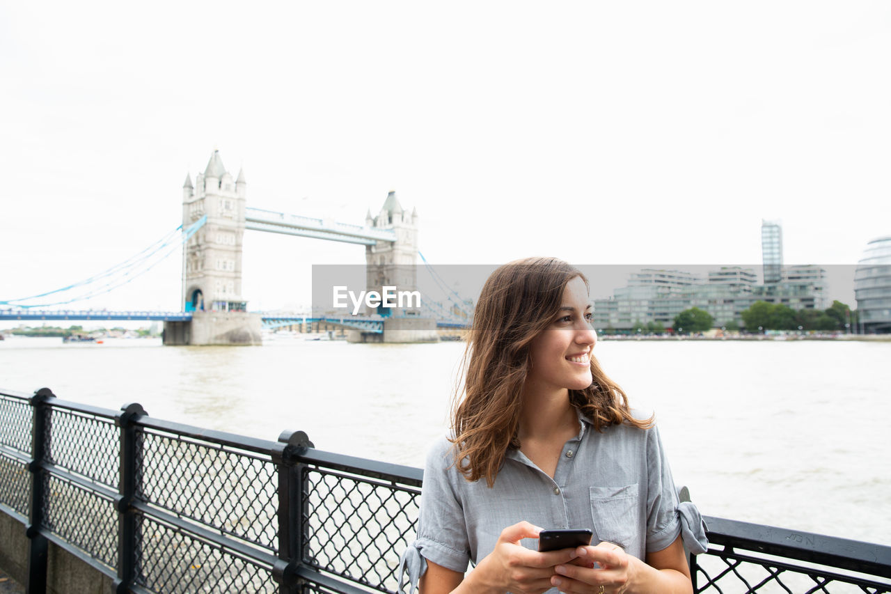 Young woman using phone by thames river with tower bridge in background