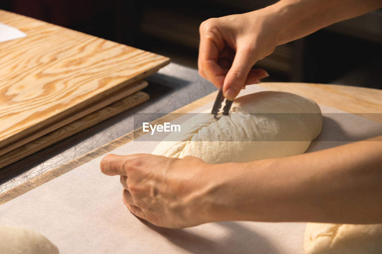 Close-up of a baker's female hands cutting a loaf of dough into a loaf of bread with a blade before