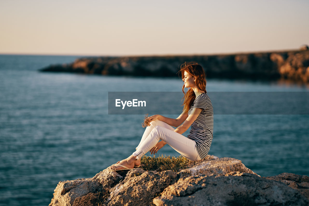 WOMAN SITTING ON ROCK BY SEA AGAINST SKY