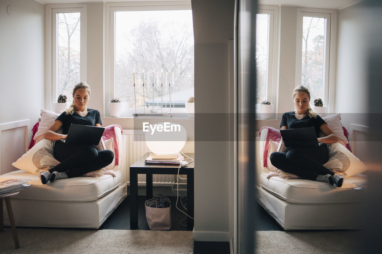 Reflection of teenage girl using laptop reclining on seat against window at home