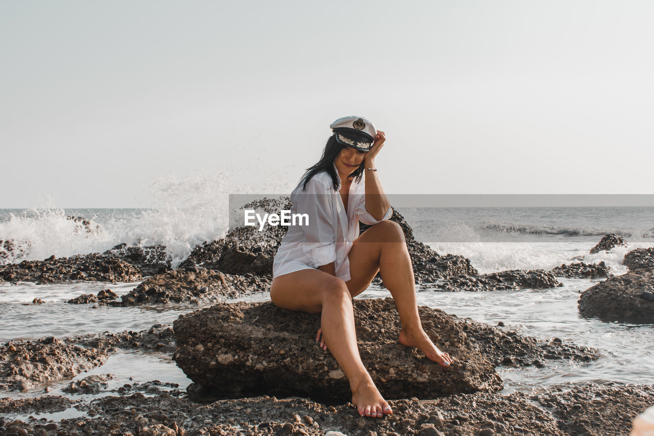 Full length of woman sitting on rock at beach against clear sky