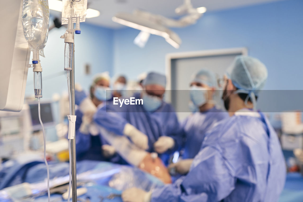 Iv drip in operation room with doctors operating surgery in background