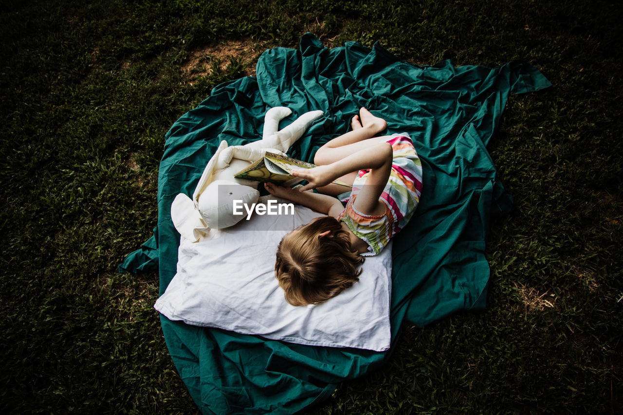 High angle view of girl reading book while lying by stuffed toy on blanket