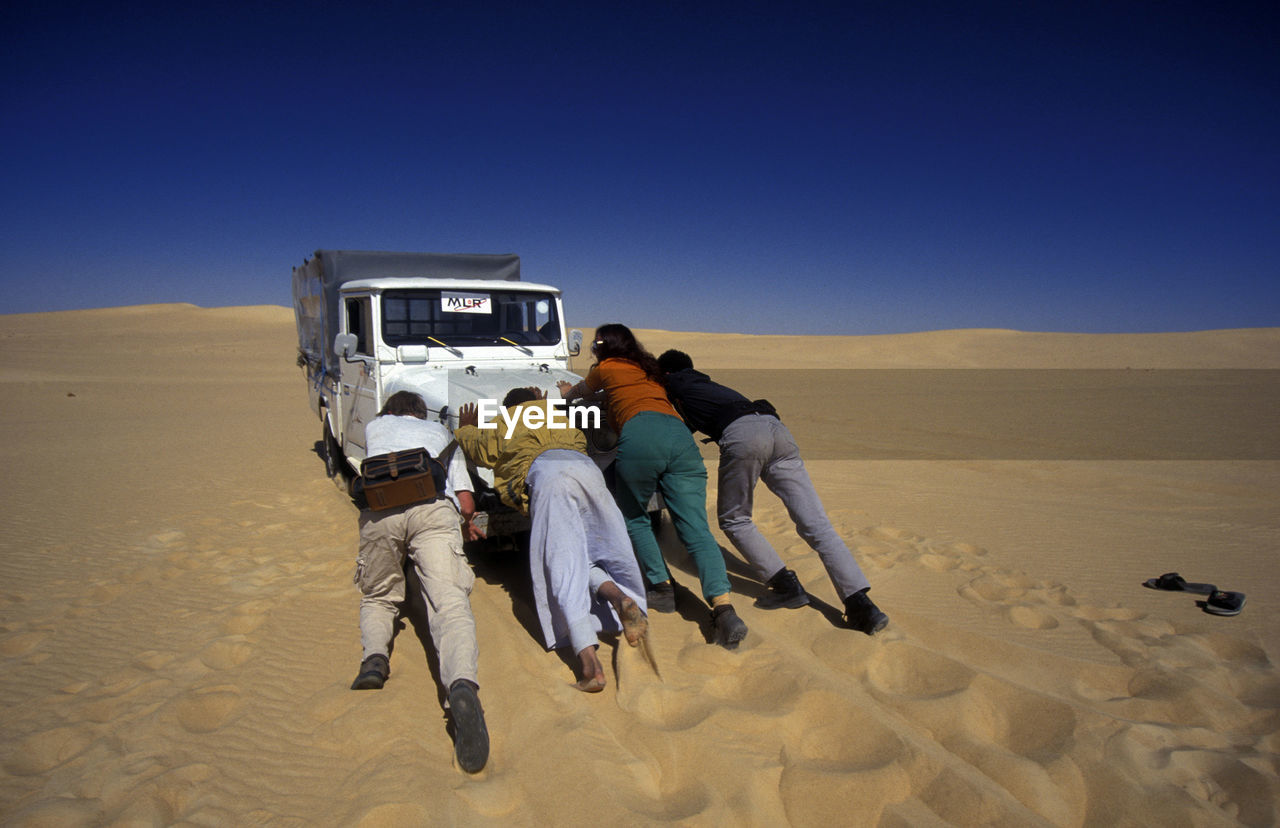 Men and women pushing jeep at desert against clear blue sky