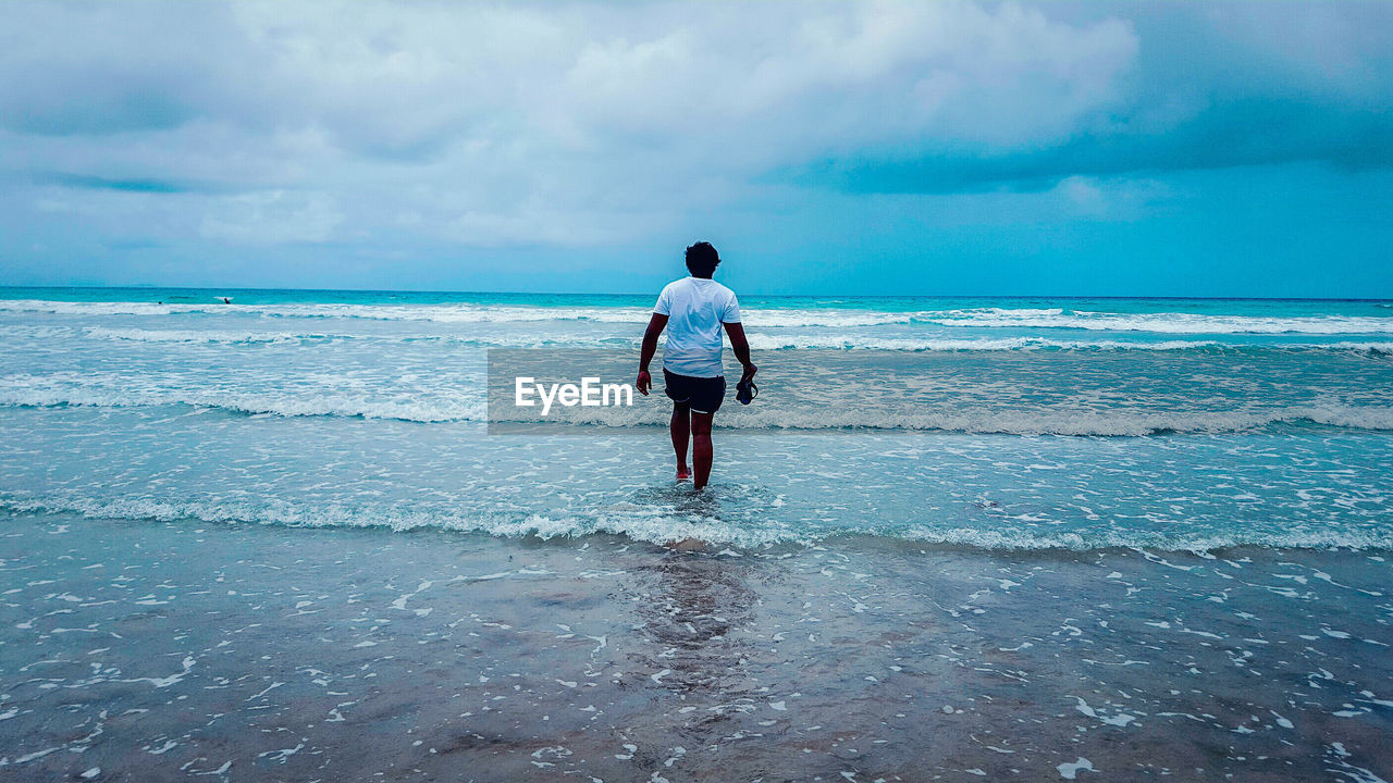 Rear view of man walking on shore at beach against cloudy sky