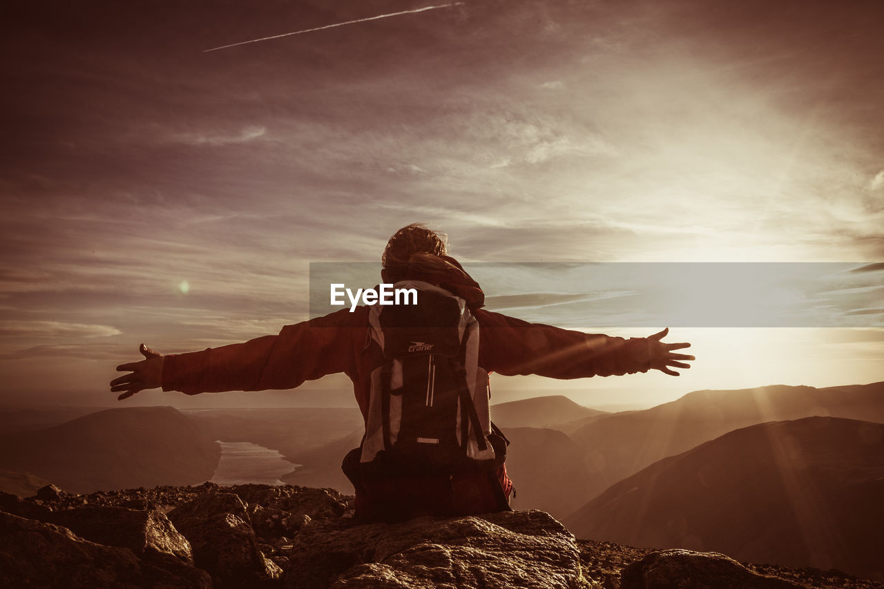 Rear view of hiker with backpack and arms outstretched leaning on rock against sky