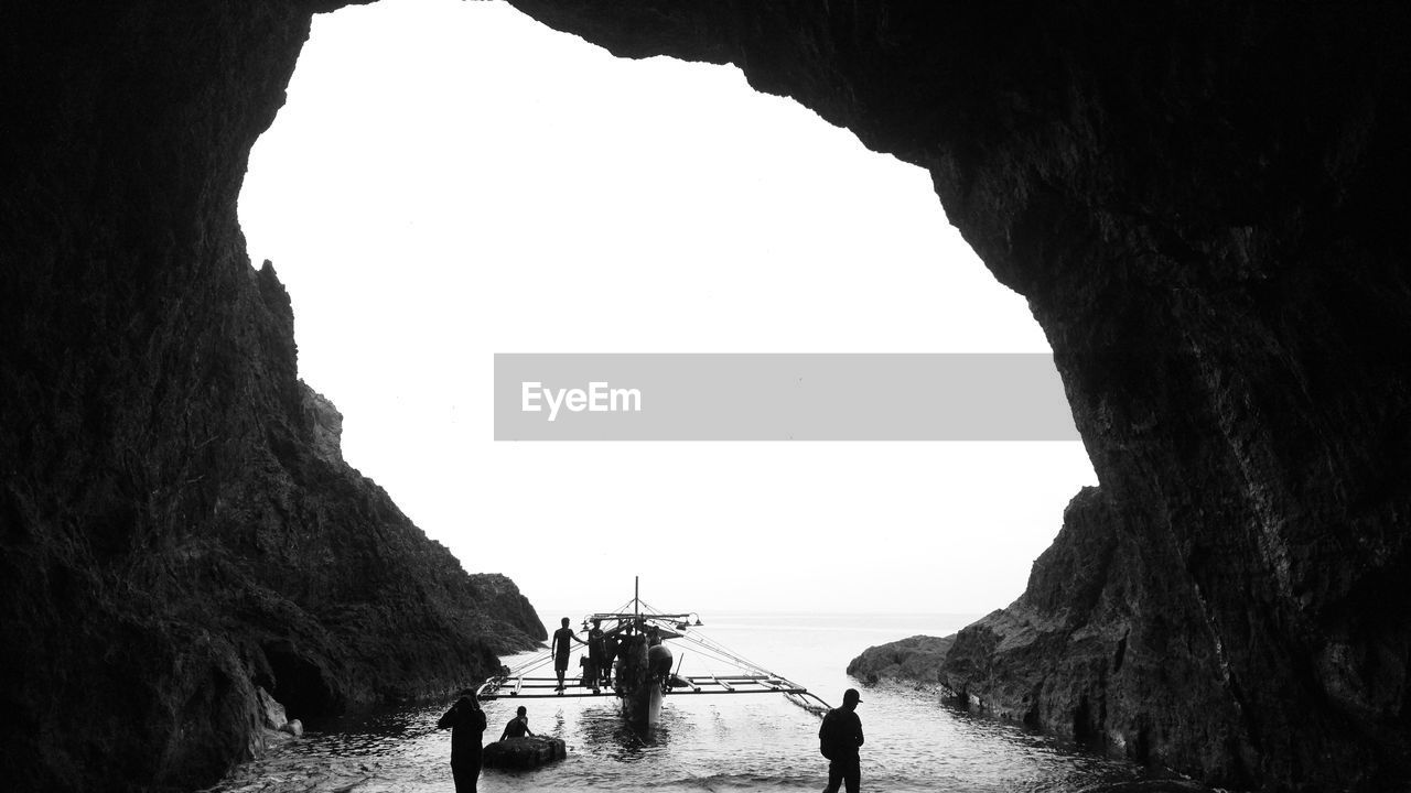 People with outrigger canoe in cave at beach against clear sky