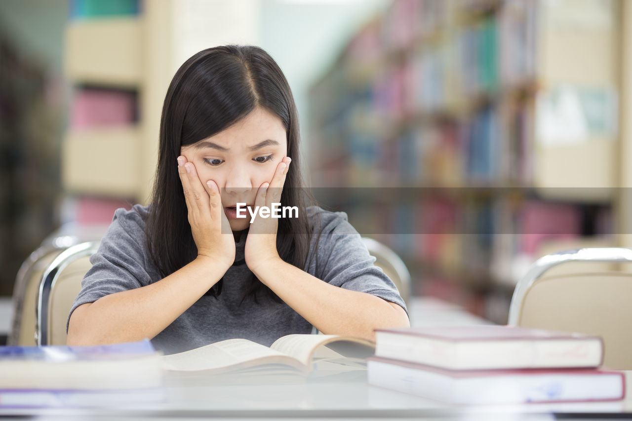 Tensed young woman reading book while sitting at table in library