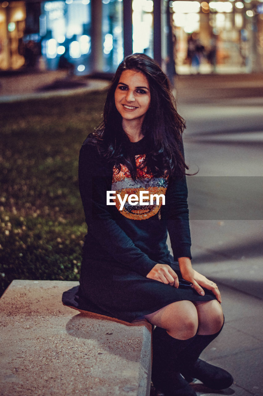 PORTRAIT OF SMILING YOUNG WOMAN SITTING ON STREET