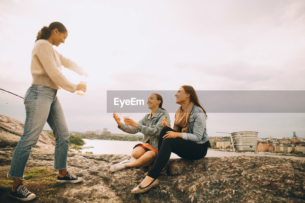 Woman giving drink to friends on rock formation at lakeshore against sky