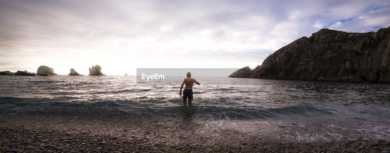 FULL LENGTH OF MAN STANDING ON ROCK AT BEACH AGAINST SKY
