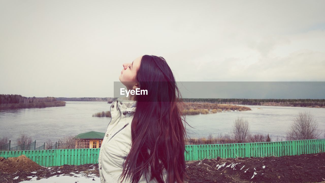 Young woman with eyes closed standing by river against sky