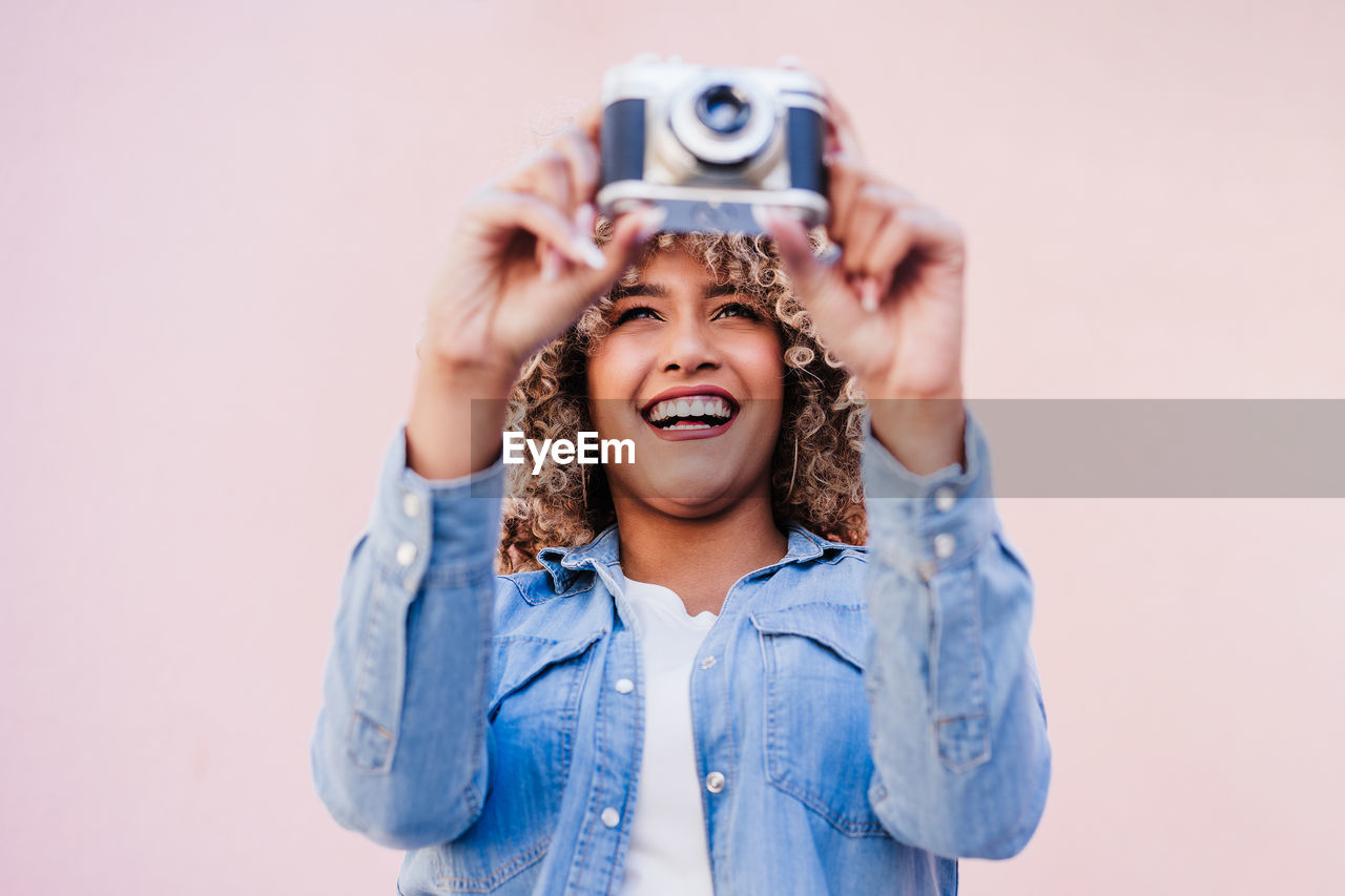 Beautiful happy hispanic woman with afro hair holding vintage camera. pink background