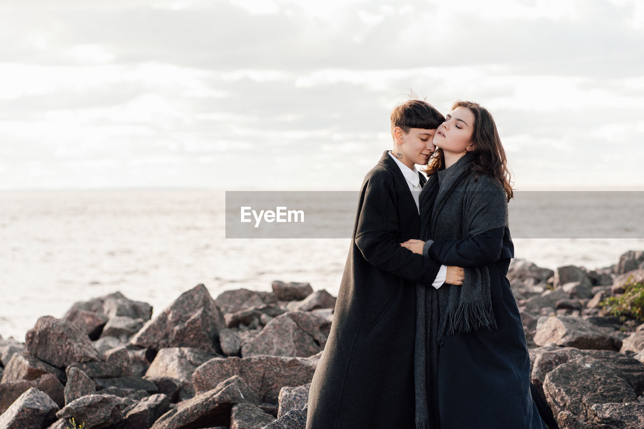 Lesbian women embracing while standing on rock against sea and sky