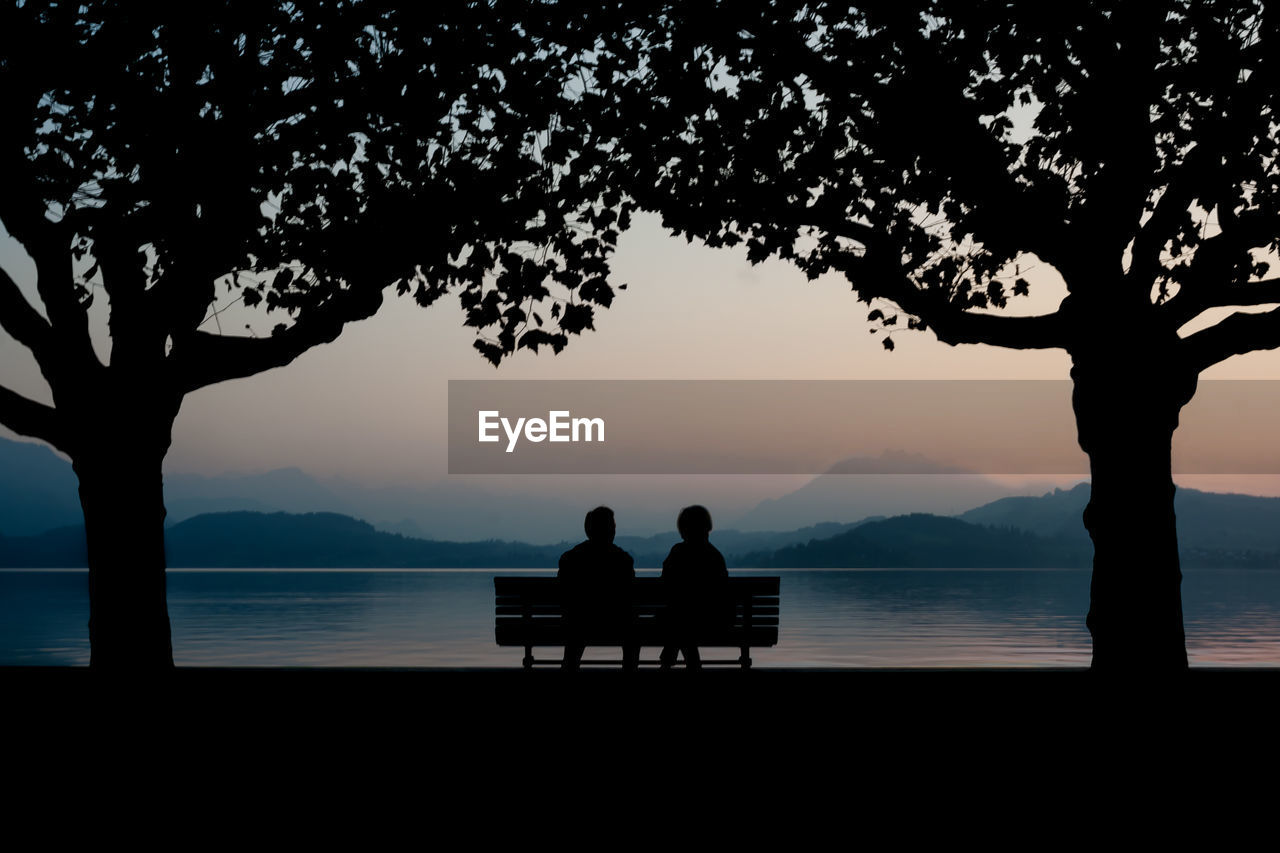 Silhouette couple sitting on bench by calm lake at dusk