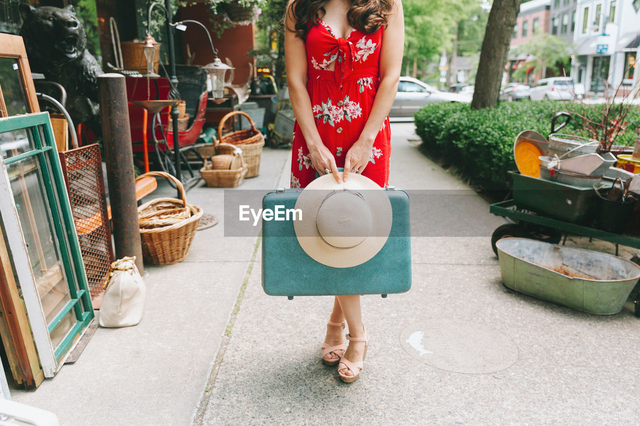 Low section of woman holding hat and briefcase while standing on footpath