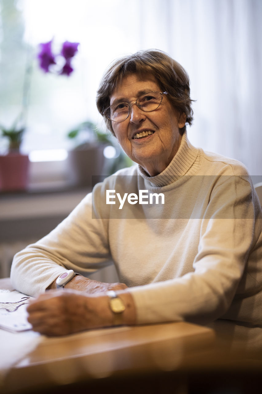 Smiling wrinkled woman with emergency button on wrist sitting by table at home