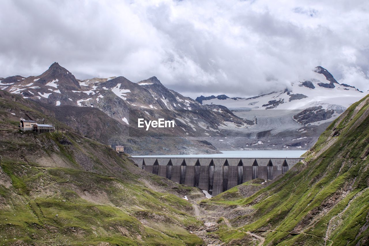 View of dam amidst snowcapped mountains against cloudy sky