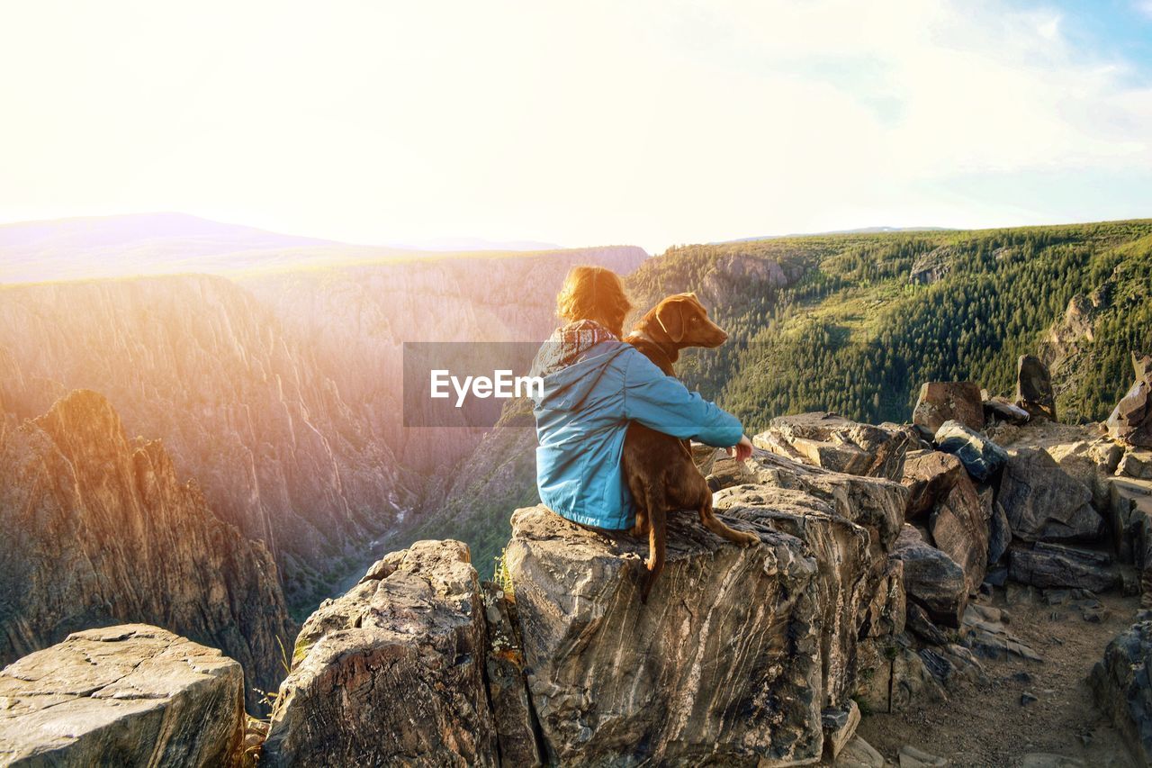 Girl sitting with dog on rocks against mountains during sunny day