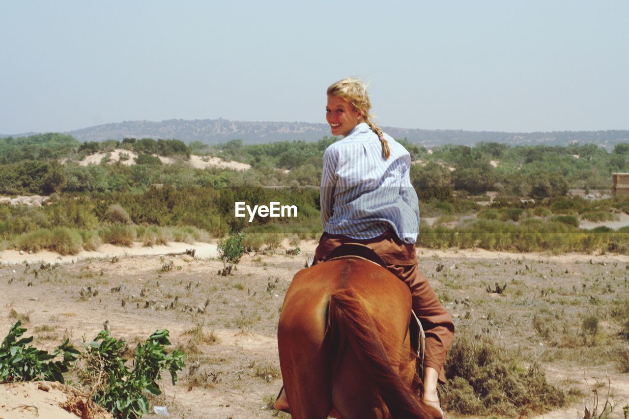 Rear view of smiling young woman riding horse against clear sky during sunny day