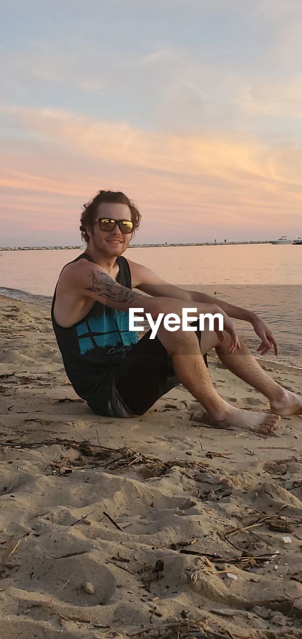 Portrait of man sitting on shore at beach against sky during sunset