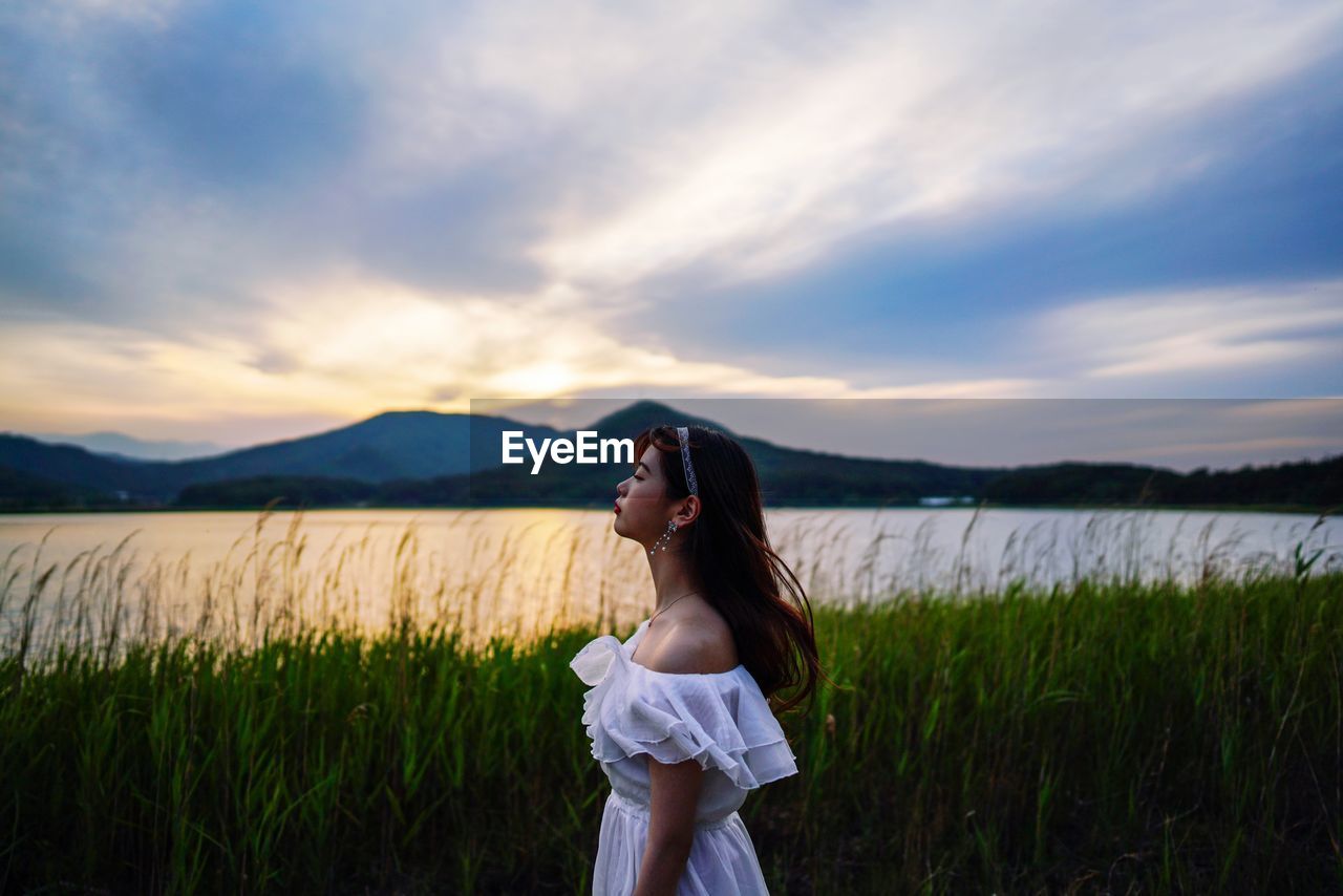 Side view of teenage girl standing on land against sky during sunset