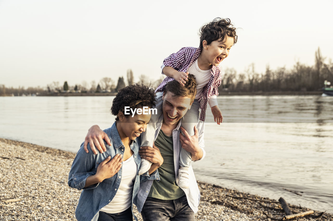 Happy man carrying boy on shoulder while walking with woman at lakeside