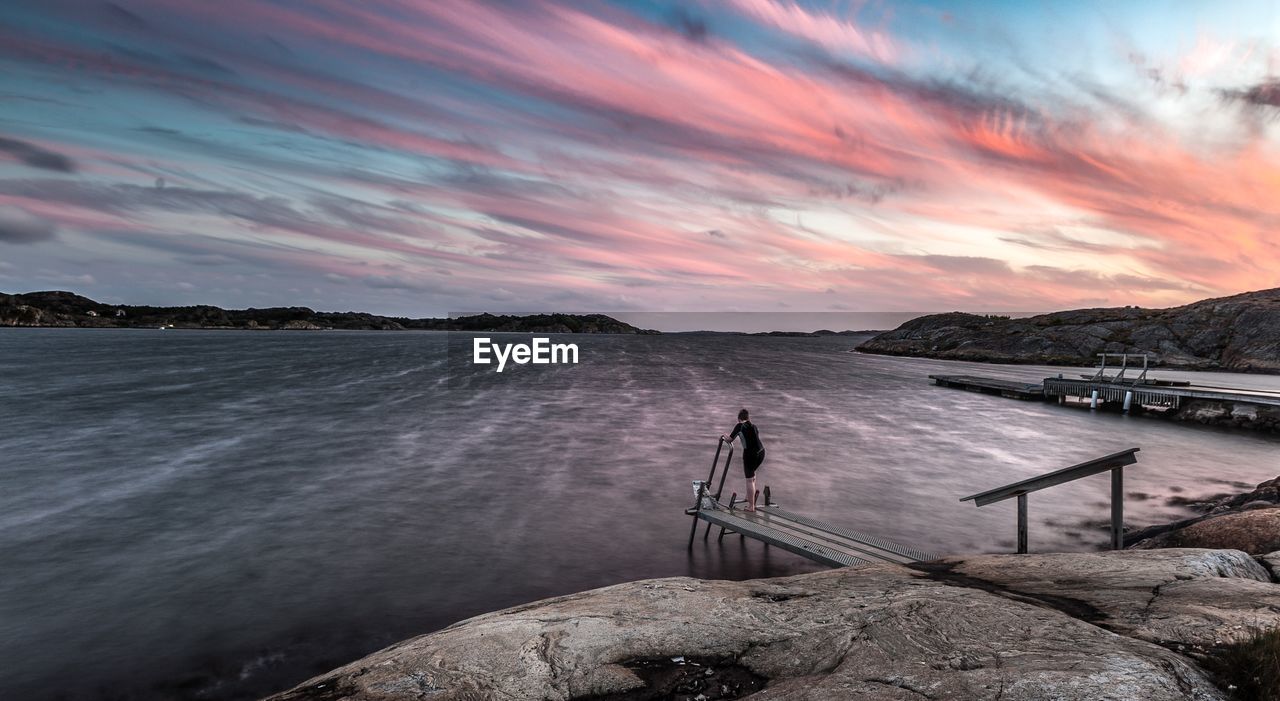 Rear view of woman on pier at lake against cloudy sky during sunset