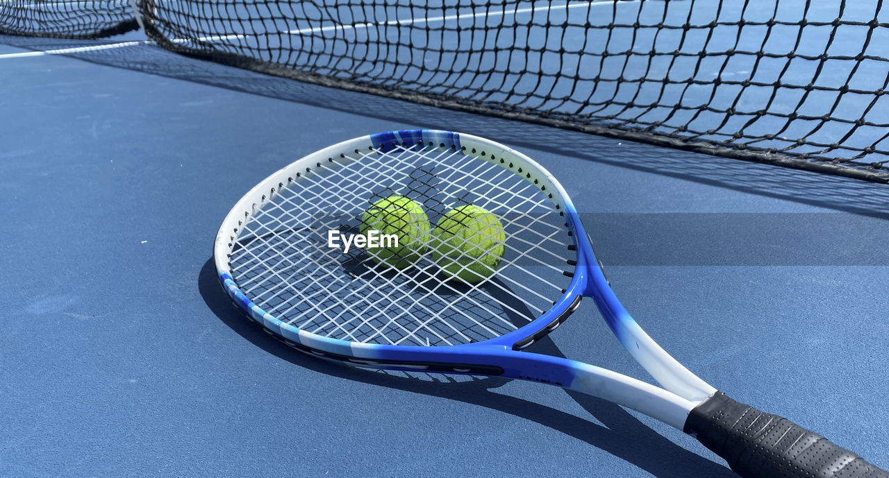 Horizontal view of two tennis balls with a racket on top on a blue tennis court.