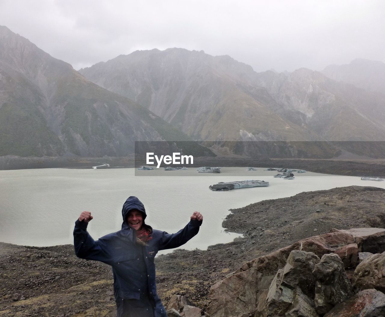 Portrait of happy hiker flexing muscle against lake and mountains during foggy weather