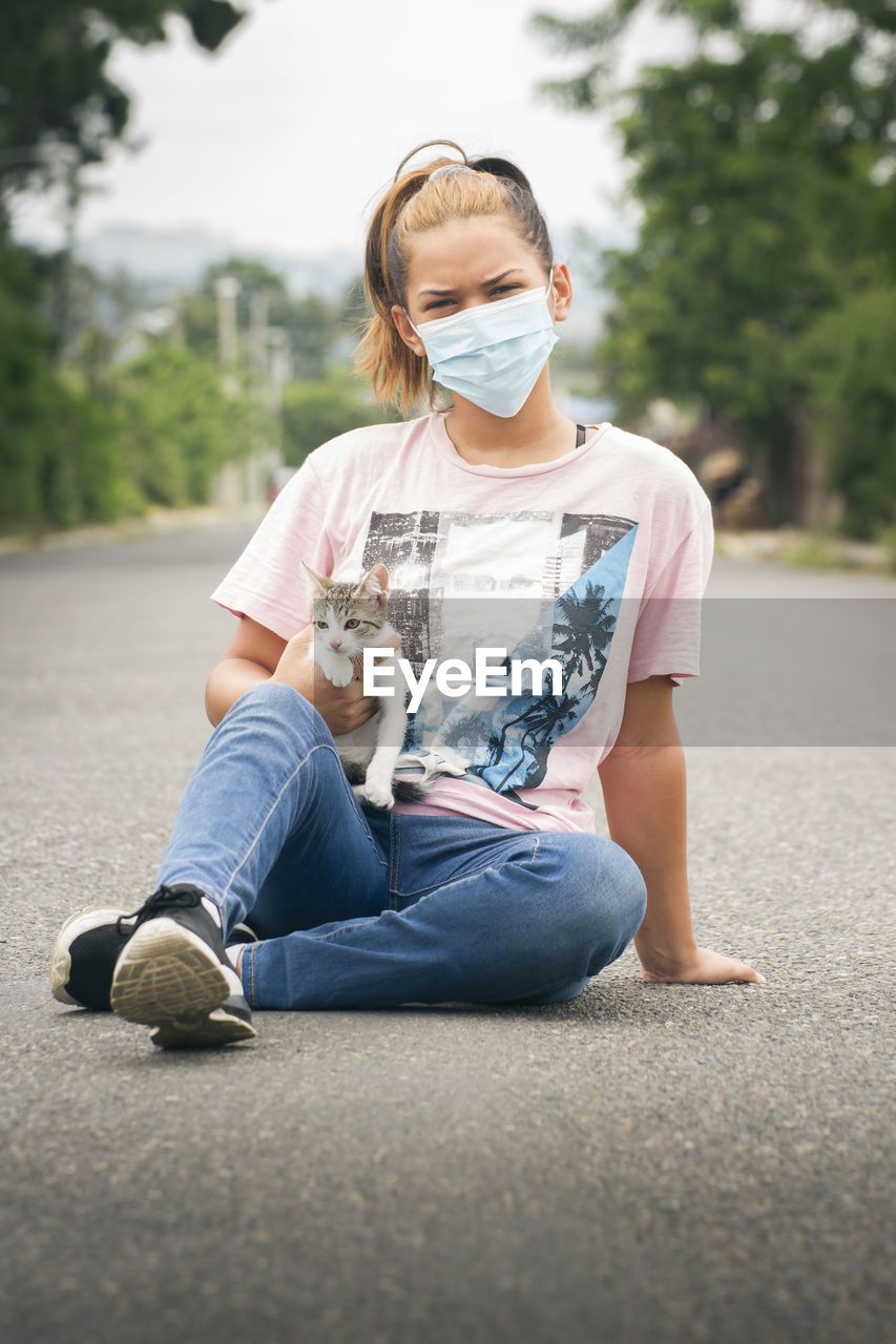 One young girl or woman wearing face mask seated on street carrying a young white and gray cat in a blurred natural environment