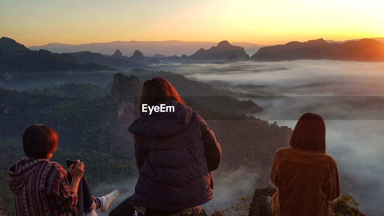 REAR VIEW OF PEOPLE LOOKING AT MOUNTAIN DURING SUNSET