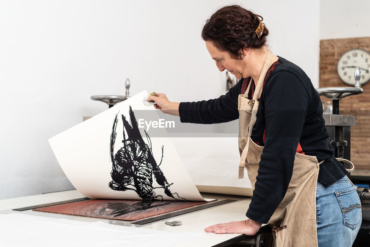 Side view of skilled female artist removing paper sheet with impression from linoleum piece placed on printing press machine while working in creative studio