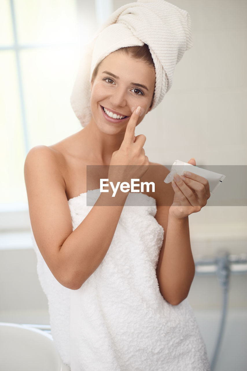 Portrait of smiling young woman applying moisturizer on face in bathroom